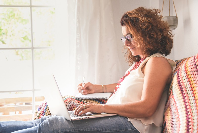 Woman sits at home reading from her laptop and taking notes