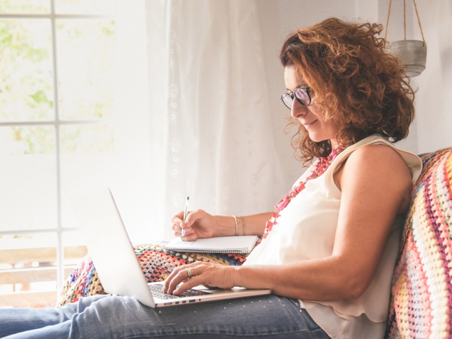 Woman sits at home reading from her laptop and taking notes